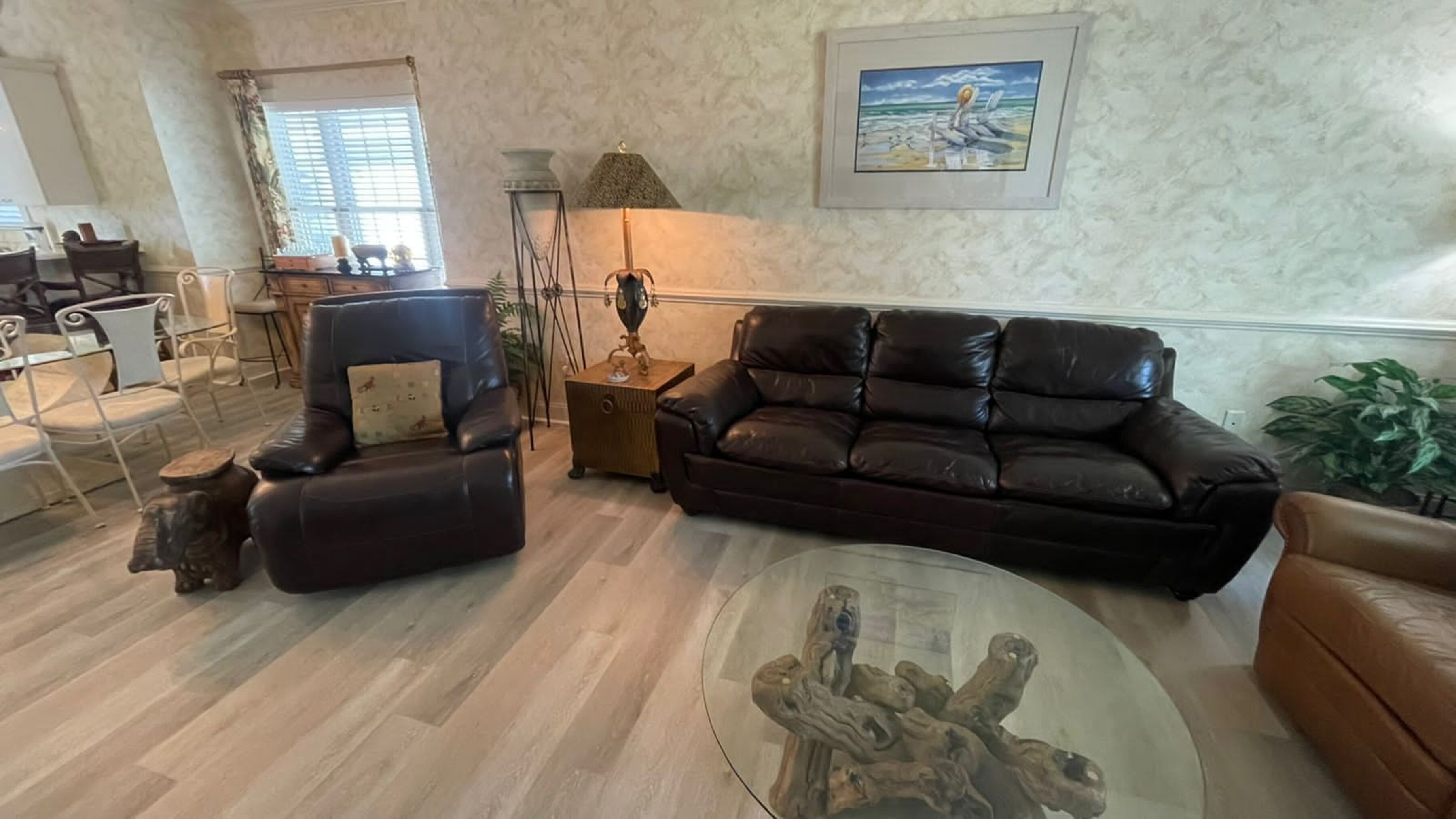 A living room with a dark brown leather sofa, a glass coffee table, and a wooden side table. There is a window with blinds, a lamp, and a framed picture on the wall.
