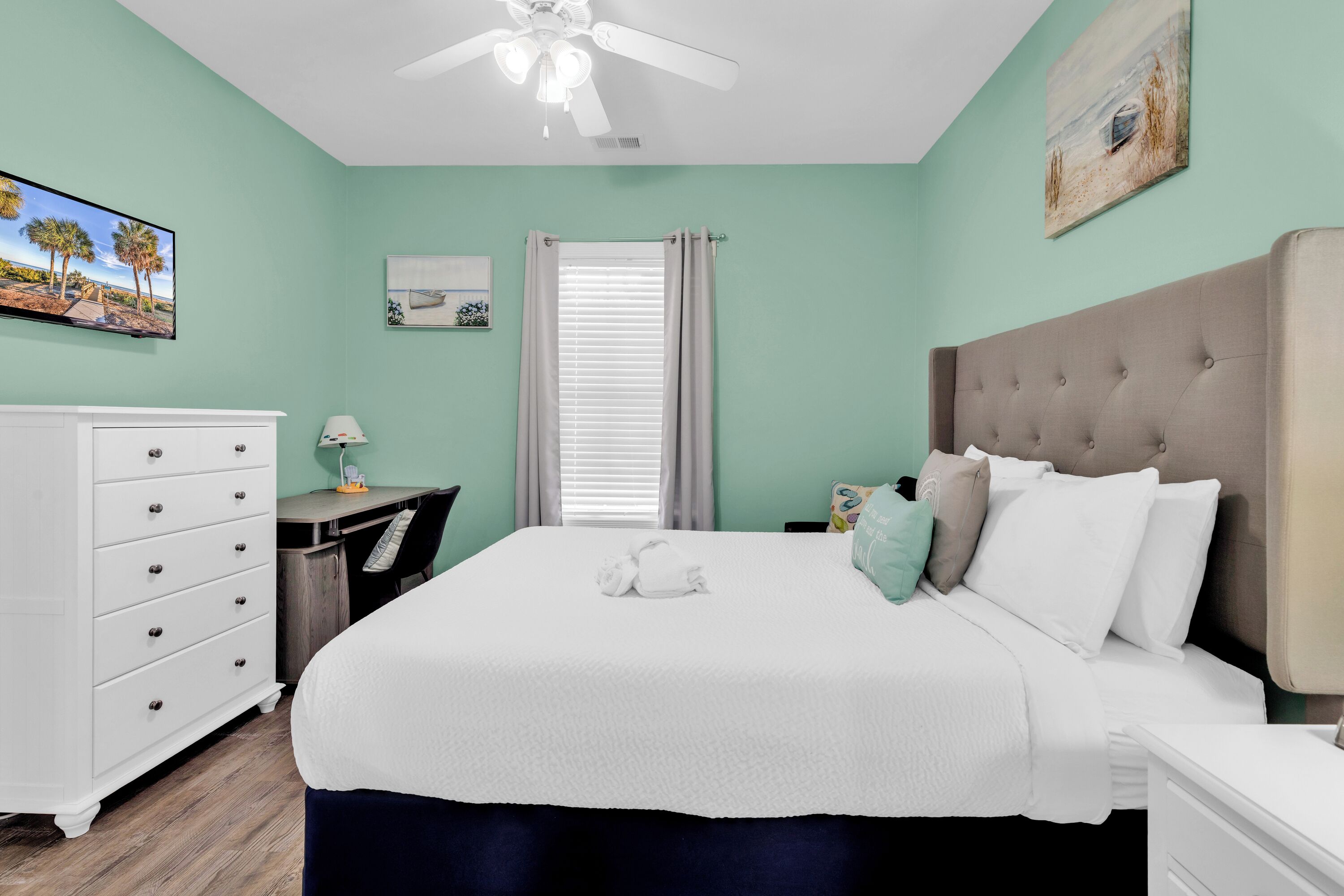 A bedroom with a bed, a desk, a chair, a dresser, a ceiling fan, and a window with curtains.