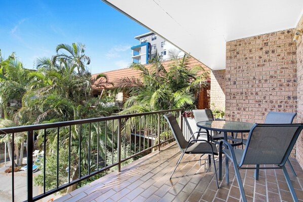 A balcony with a glass table and chairs, a brick wall, and a view of a building and palm trees.