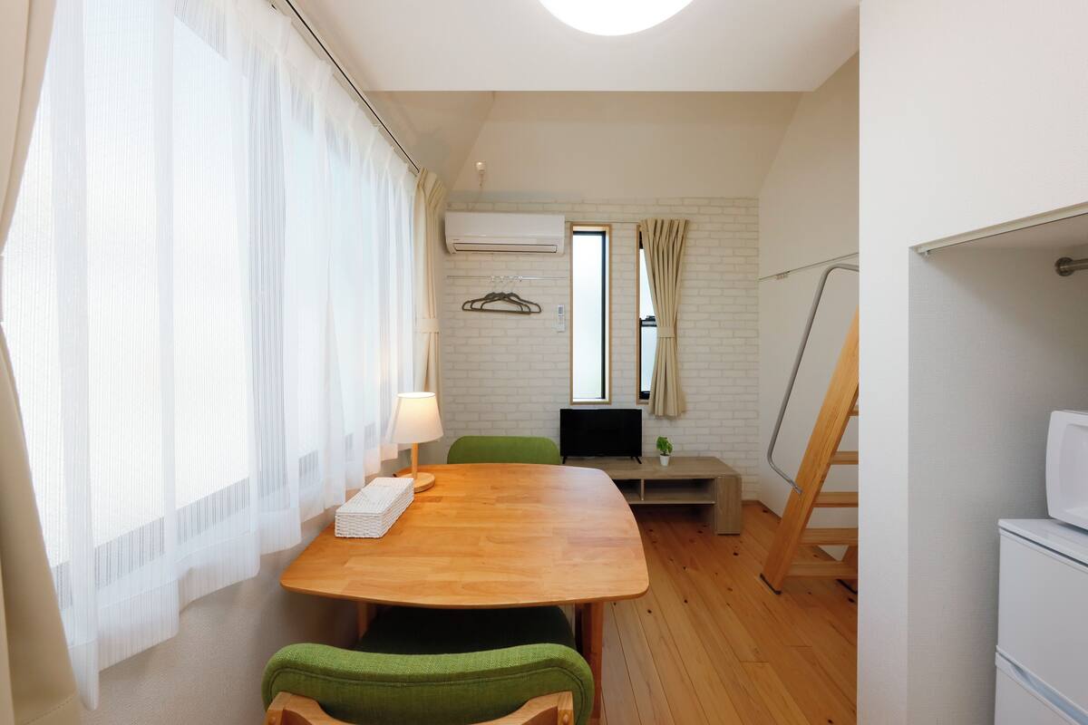 A small, well-lit room with a wooden table, green chairs, a TV, and a white air conditioning unit on the wall.