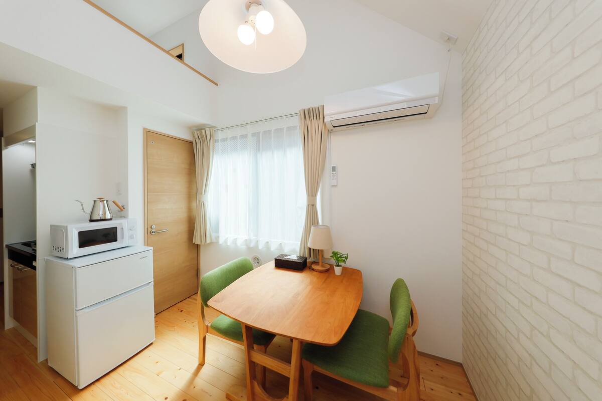 A compact kitchen with a microwave, a small refrigerator, and a wooden dining table with green chairs.