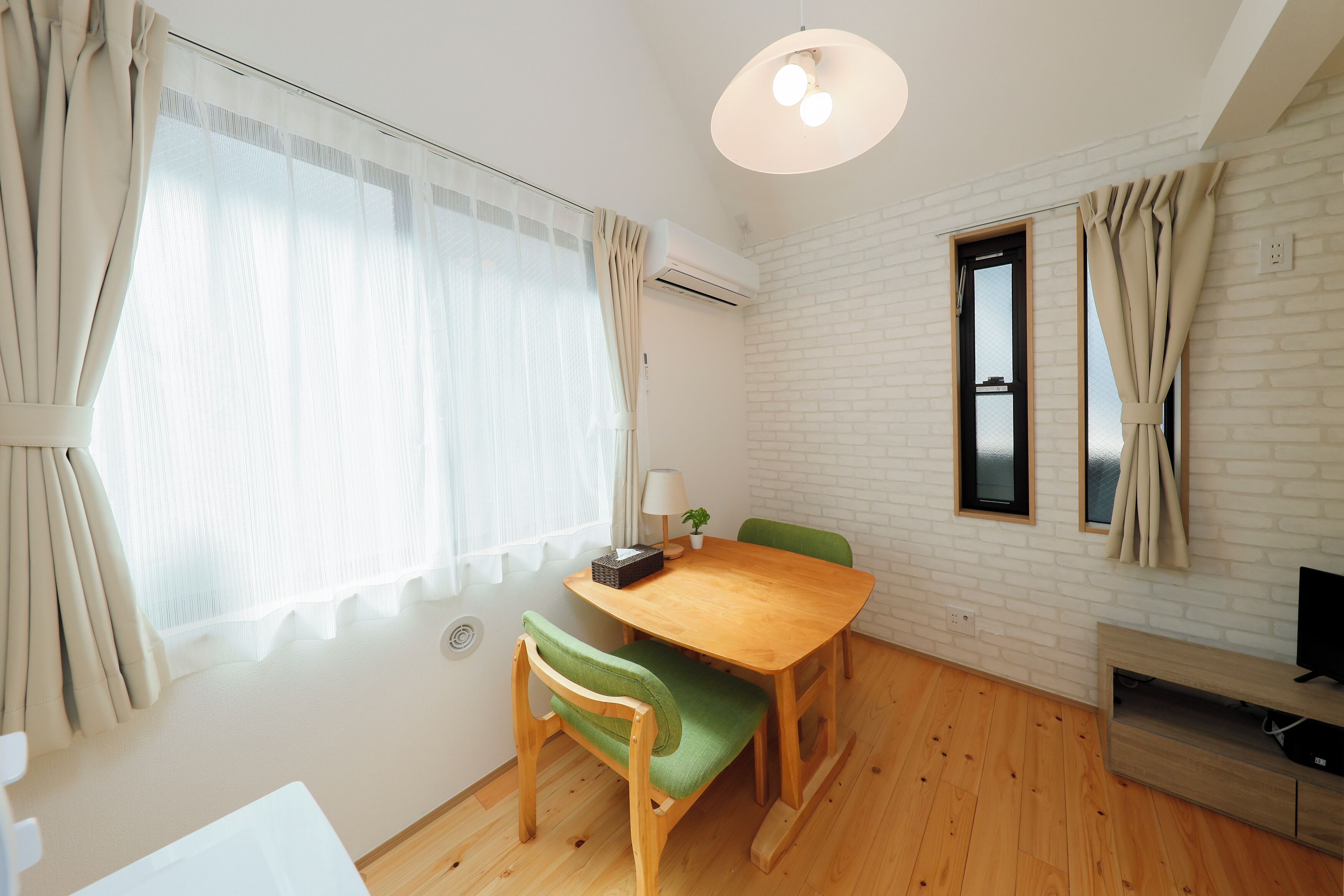 Una habitación con una mesa de comedor de madera, sillas con acolchado verde, un escritorio blanco, una planta pequeña y un televisor sobre un mueble de madera.