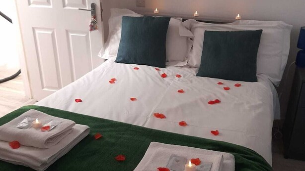 A neatly made bed with white linens, green pillows, and rose petals scattered across the bedspread. There are two lit candles on the bed, and a stack of folded towels with a lit candle on top. - The Broadway Rooms  (Leicester)