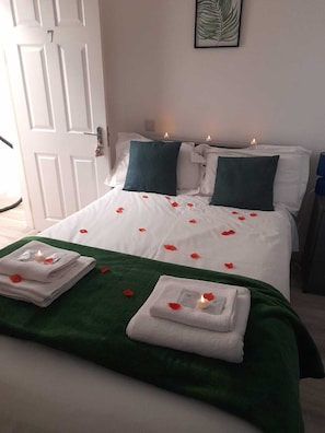 A neatly made bed with white linens, green pillows, and rose petals scattered across the bedspread. There are two lit candles on the bed, and a stack of folded towels with a lit candle on top. - The Broadway Rooms  (Leicester)