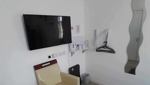 A small, modern room with a flat-screen TV mounted on the wall, a beige armchair, a small shelf with cups and a hook, and a patterned rug on the floor. - The Broadway Rooms  (Leicester)