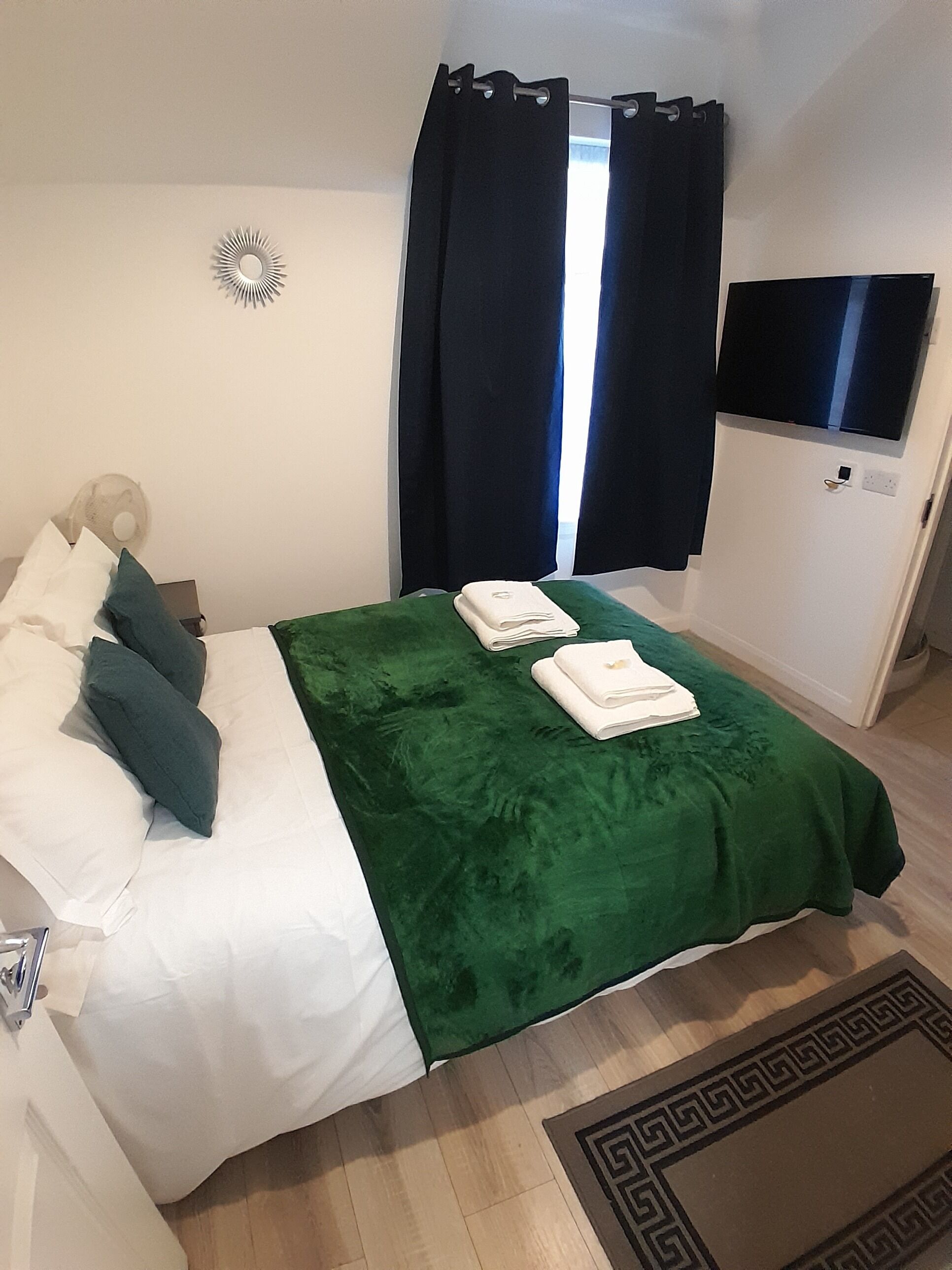 A neatly made bed with a green blanket, white pillows, and folded towels. There is a small decorative item on the wall, a dark curtain covering a window, and a flat-screen TV mounted on the wall.
