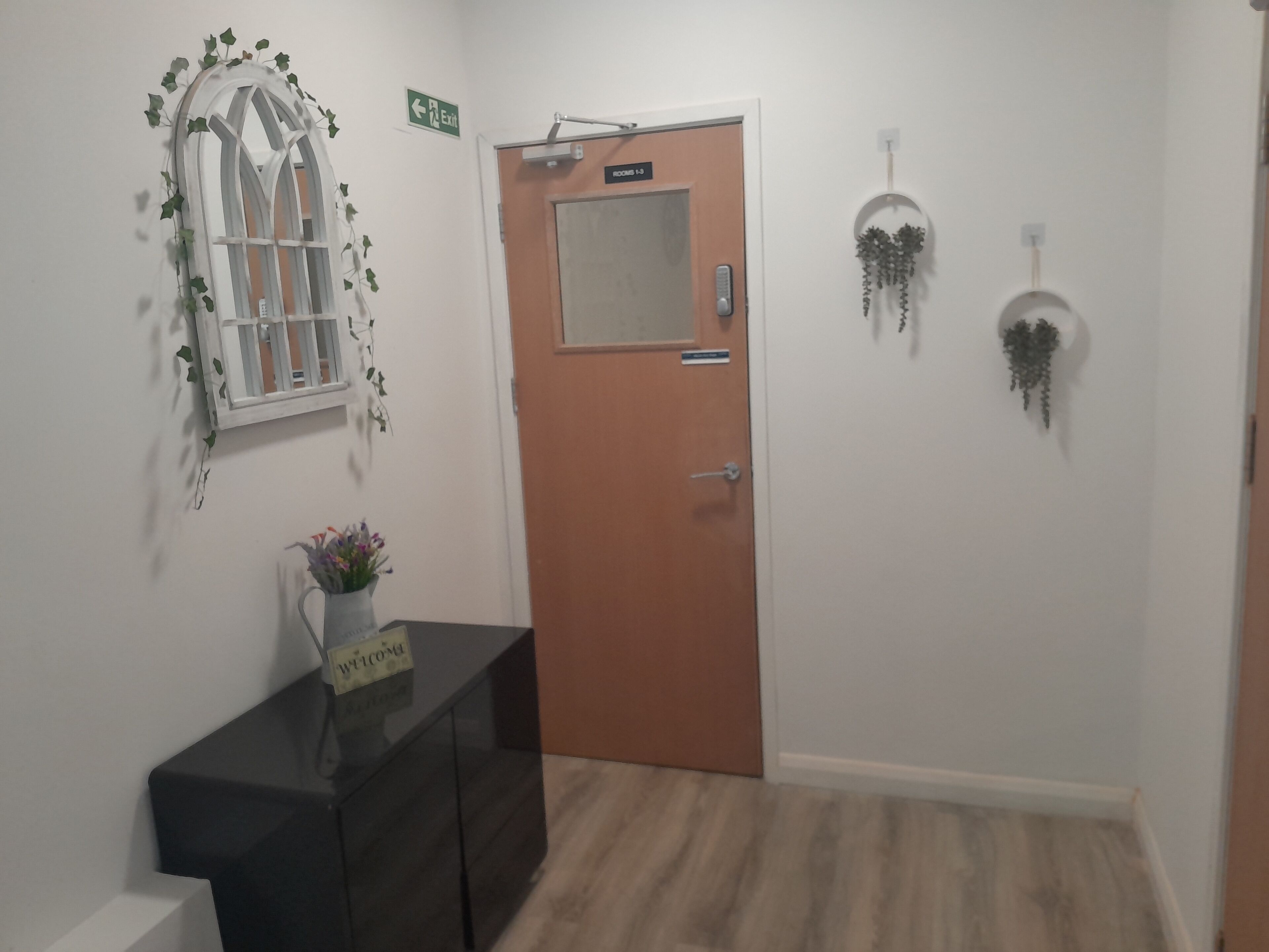 A hallway with a wooden door labeled "ROOM 1-3", a small table with a vase of flowers, and wall-mounted decorations including a mirror and hanging plants.