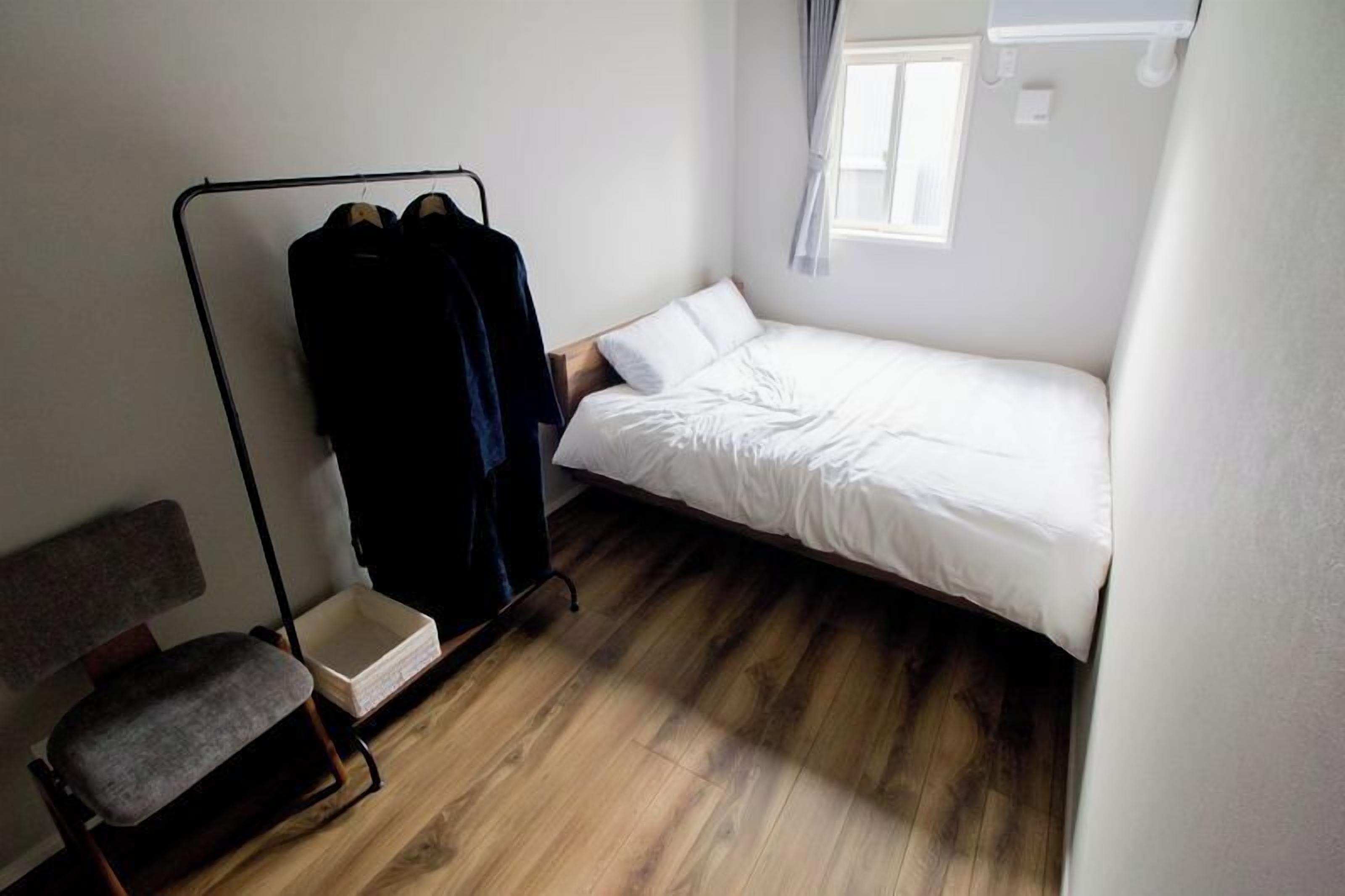 A small, single-bed room with a wooden floor, a chair, a coat rack with clothes, and a window with curtains.