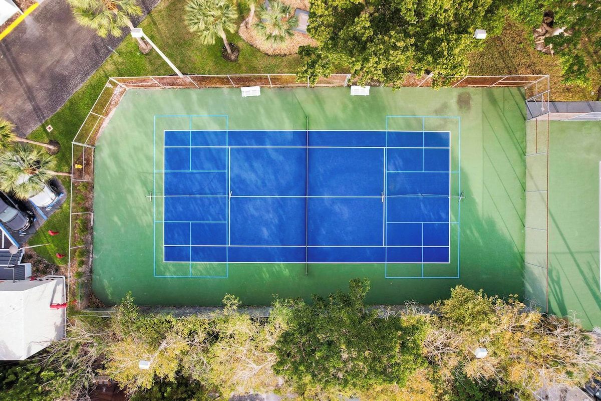 An aerial view of a tennis court with surrounding greenery and a building in the background.