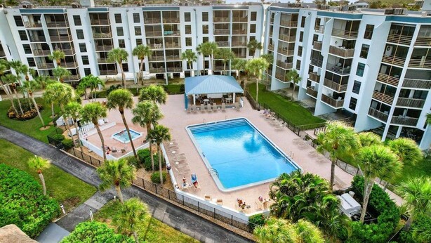 Ein mehrstöckiges Hotel mit einem zentral gelegenen Swimmingpool, umgeben von Palmen und Liegestühlen.