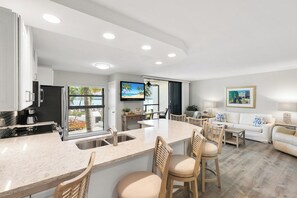 A modern kitchen with a central island, bar stools, and a dining area with a sofa and chairs. - The Riverside Club - B206 (Marco Island)