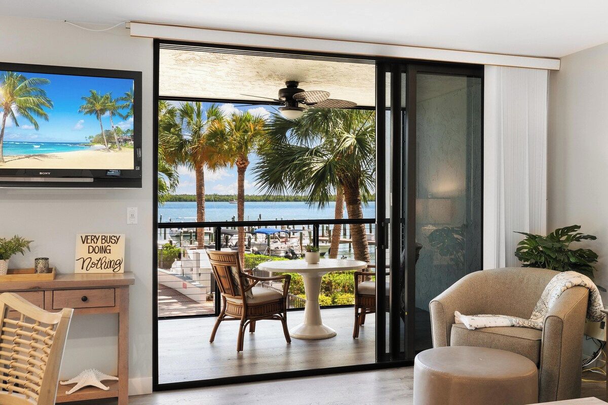 A room with a large sliding glass door leading to a balcony with a view of palm trees and the sea.