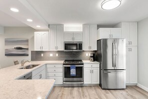 A modern kitchen with white cabinets, stainless steel appliances, and a grey backsplash. - The Riverside Club - B206 (Marco Island)