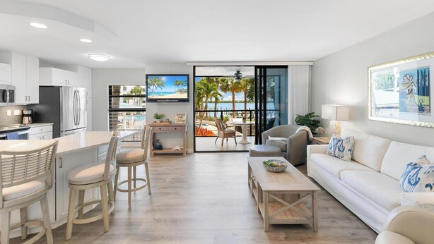 A modern kitchen with white cabinets, a central island with bar stools, and a dining area with a view of the beach. - The Riverside Club - B206 (Marco Island)