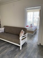 Una sala de estar moderna con un sofá marrón y un sillón blanco, una mesita con un cojín decorativo, y vistas a un balcón con barandilla blanca y una ventana con cortinas de encaje.