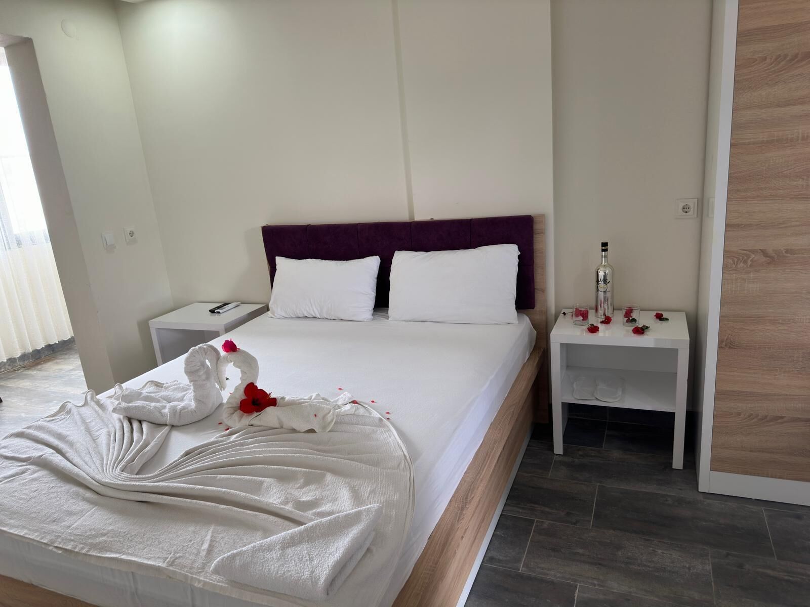 A neatly made bed with white linens and a purple headboard, a bedside table with a bottle and glasses, and a small table with red flowers.