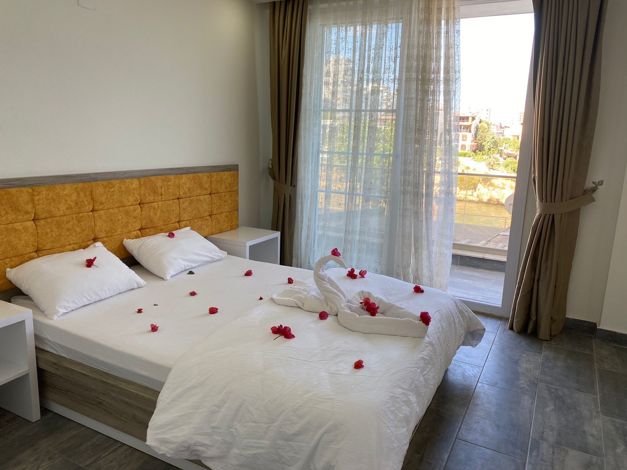 Una habitación de hotel con cama, almohadas blancas, cabecero de madera, mesita de noche y vistas al exterior a través de una ventana con cortinas.