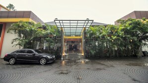A building entrance with a glass canopy, a black car parked in front, and a paved area with palm trees. - Collection O 295 Grha Ciumbuleuit Guest House (Bandung)