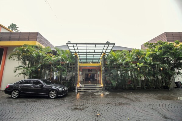 A building entrance with a glass canopy, a black car parked in front, and a paved area with palm trees.