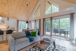 A spacious living area with a high ceiling, large windows, a sofa, a dining table, and a kitchen area.