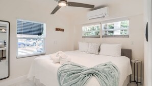 A bedroom with a bed, a ceiling fan, an air conditioning unit, and two windows with blinds.
