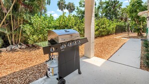 An outdoor grill area with a gas tank, under a wooden pergola, surrounded by greenery and a paved path.