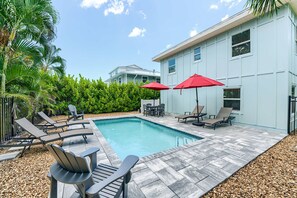 A swimming pool with lounge chairs and umbrellas, surrounded by a hedge and a building.