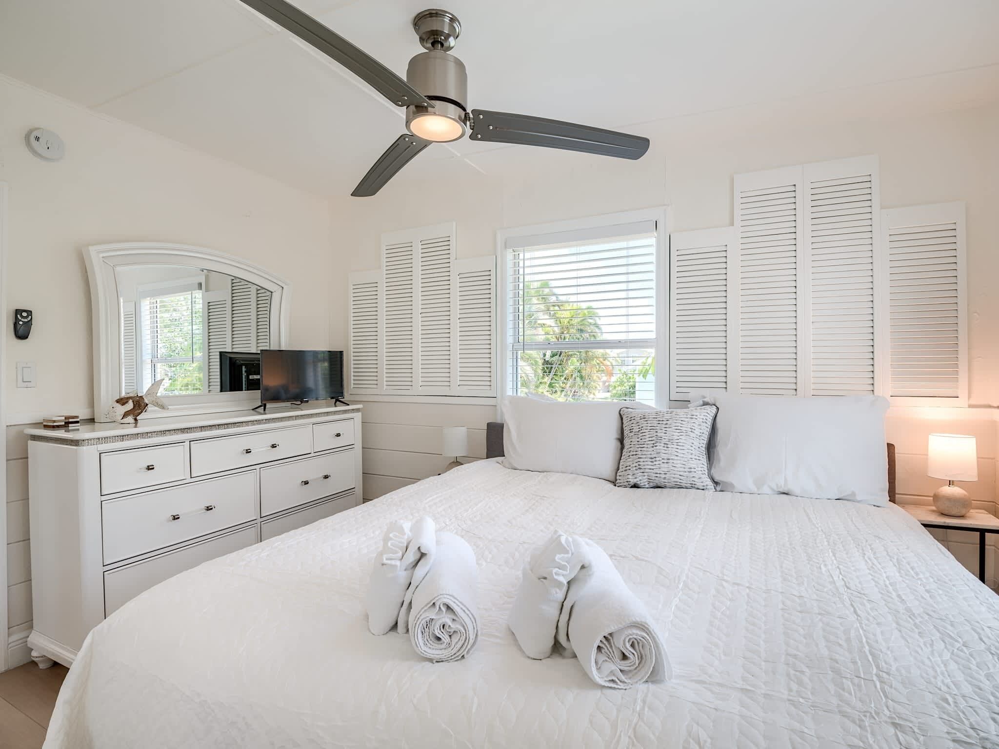 Un dormitorio con cama, ventilador de techo, cómoda, televisor y ventana con contraventanas.