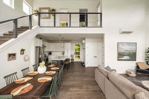 Ett rymligt vardagsrum med högt tak, en trappa, ett matbord för fyra personer och en stor soffa.