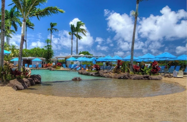 A resort pool area with palm trees, lounge chairs, and blue umbrellas.