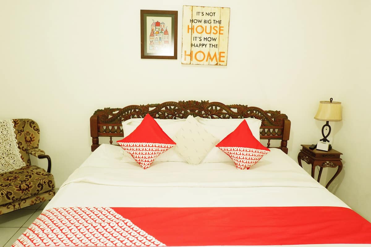 A neatly made bed with a red and white patterned bedspread, two decorative pillows, a wooden headboard, a framed picture on the wall, and a small bedside table with a lamp.