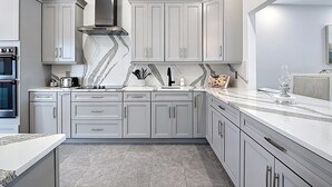 A modern kitchen with grey cabinets, a stainless steel range hood, a built-in oven, and a marble countertop. - Wally's Fairway To Haven: Discover Your Paradise: Rental with Lake View, Pool, and Golf Access! (Cape Coral)