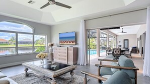 A spacious living room with a large window, a ceiling fan, a flat-screen TV, and a view of a swimming pool and outdoor seating area. - Wally's Fairway To Haven: Discover Your Paradise: Rental with Lake View, Pool, and Golf Access! (Cape Coral)