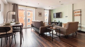 A modern living room with a sofa, a dining table set with plates and glasses, a flat-screen TV mounted on the wall, and a large window with curtains.