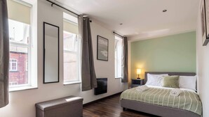 A bedroom with a bed, a bench, a bedside table, a wall-mounted TV, and a window with curtains.