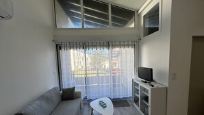 Un salon moderne avec un canapé, une petite table, un téléviseur posé sur un meuble et de grandes fenêtres avec des rideaux.