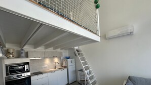 Une cuisine compacte équipée d’un four à micro-ondes, d’une machine à café et d’un évier. Une échelle est adossée au mur, et un canapé blanc est visible au premier plan.