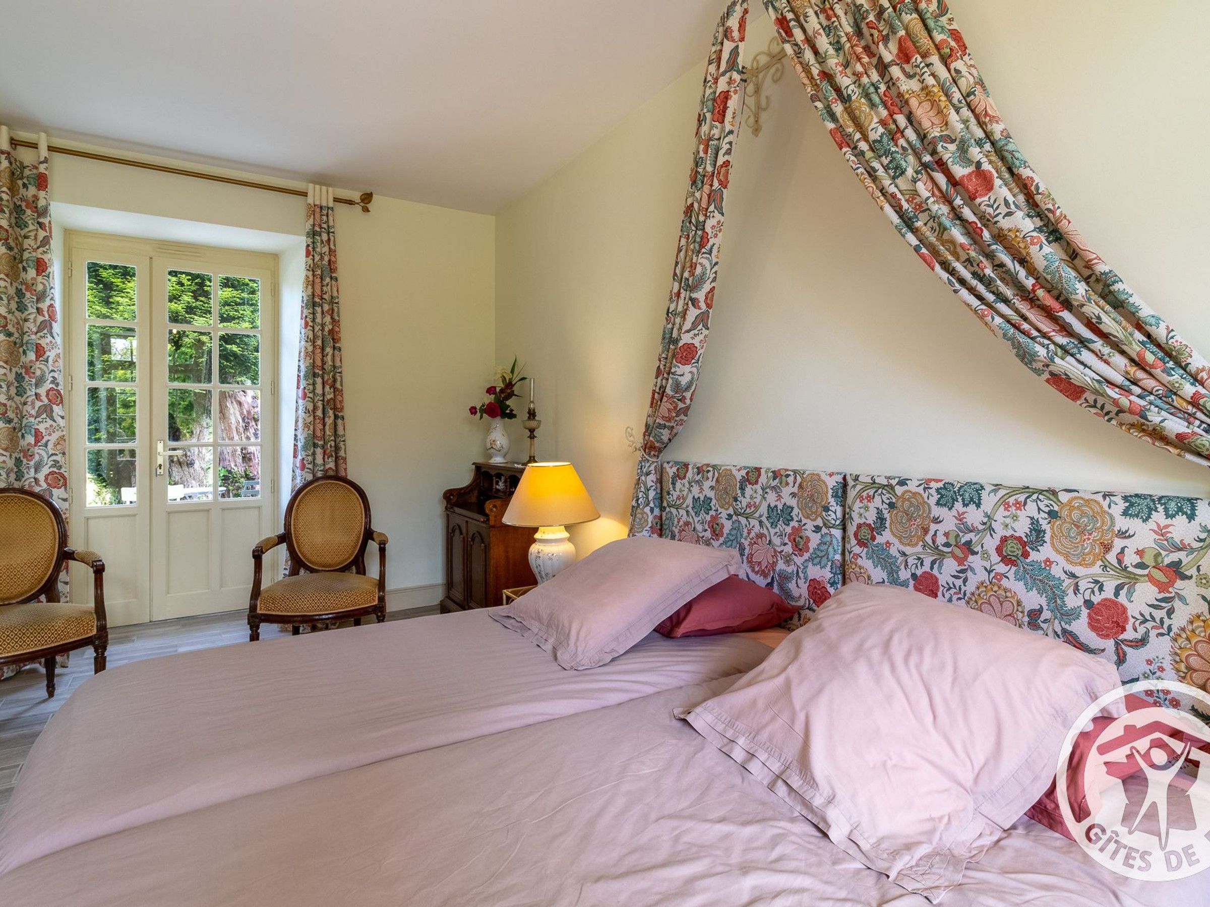 A bedroom with a floral-patterned headboard, a bed with pink bedding, two wicker chairs, a wooden nightstand with a lamp, and a window with curtains.