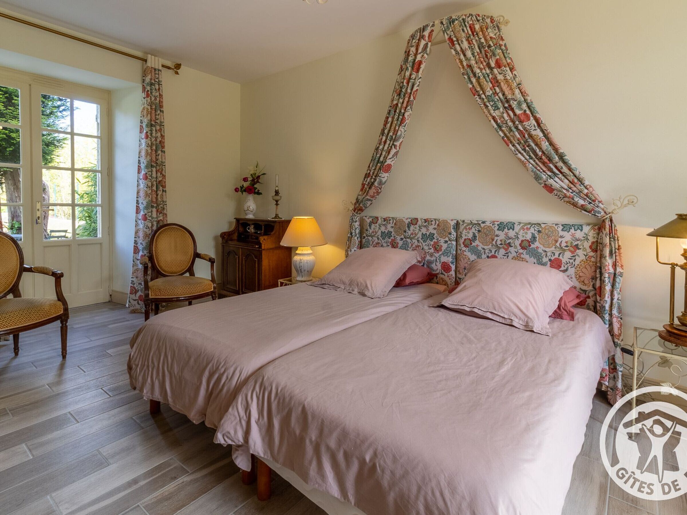 Une chambre à coucher avec un lit double, des rideaux à motifs floraux, une table de chevet avec une lampe et une chaise.