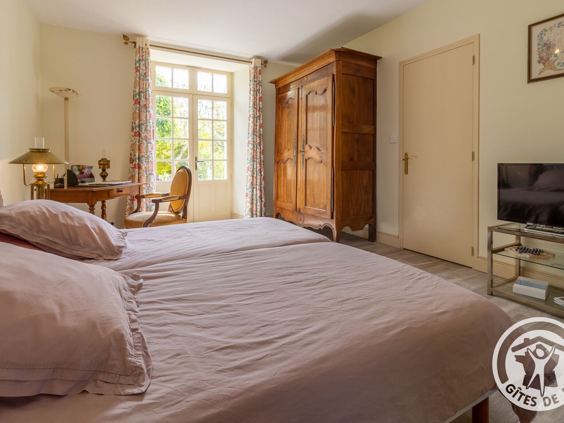 Une chambre à coucher avec un grand lit, une armoire en bois, un bureau avec une lampe et un téléviseur posé sur un meuble en verre.