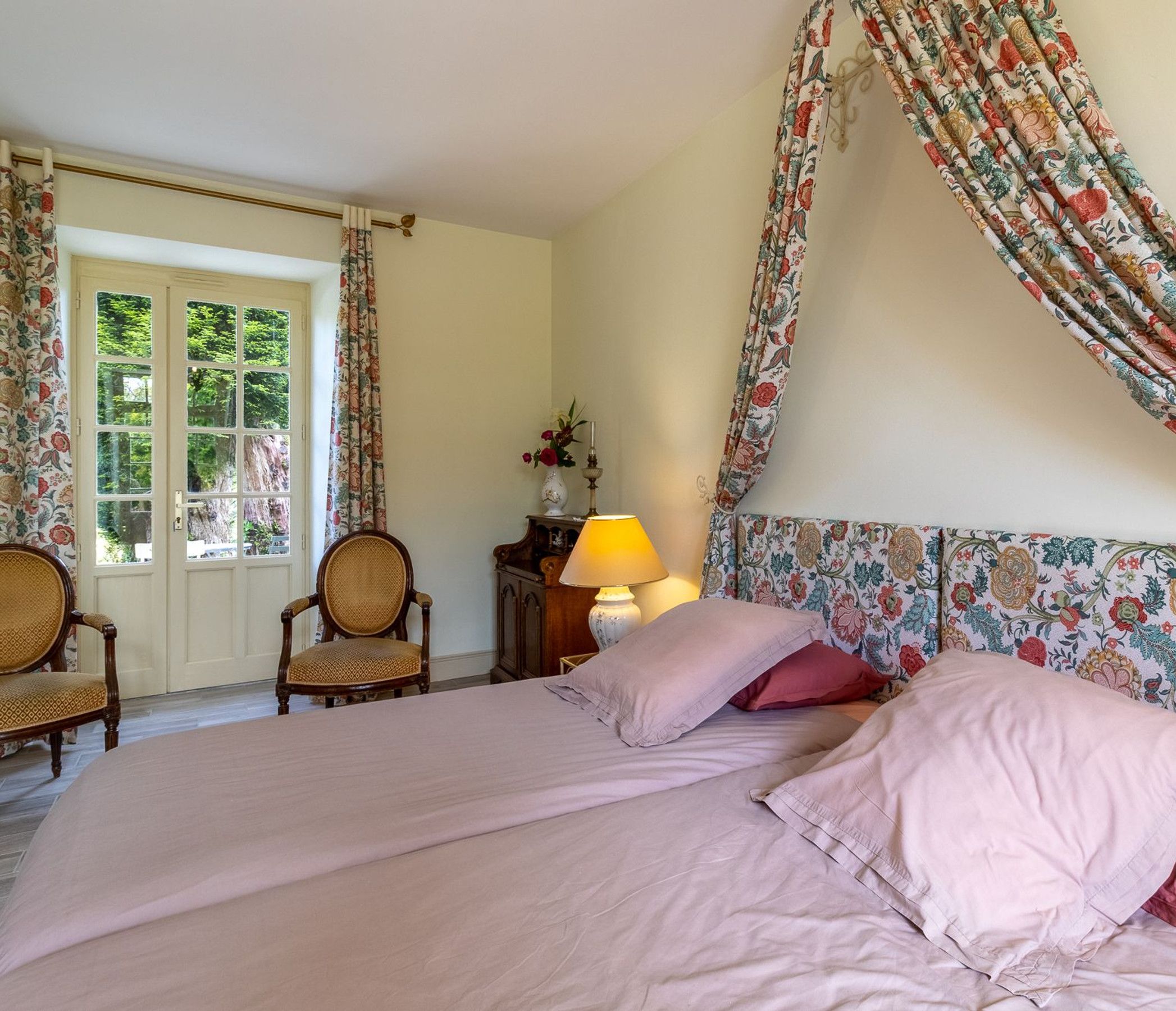 Ein Schlafzimmer mit einem blumengemusterten Kopfteil, einem Bett mit rosa Bettwäsche, zwei Korbstühlen, einem Nachttisch aus Holz mit einer Lampe und einem Fenster mit Vorhängen.