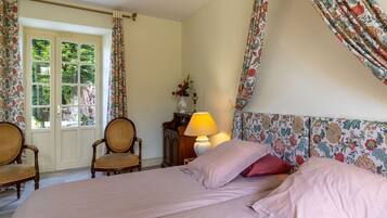 Ein Schlafzimmer mit einem blumengemusterten Kopfteil, einem Bett mit rosa Bettwäsche, zwei Korbstühlen, einem Nachttisch aus Holz mit einer Lampe und einem Fenster mit Vorhängen.