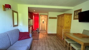 Ein Wohnzimmer mit grauem Sofa, einem Holzesstisch mit Stühlen, einem an der Wand befestigten Fernseher und einer durch einen offenen Durchgang sichtbaren Küche.