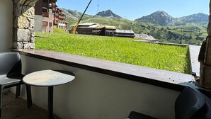 A balcony with a stone arch, a small table, and two chairs, overlooking a grassy area and mountains. - 3BR Apartment · 70m² · Ski-in/Ski-out · Pool · Pets allowed (Belle Plagne)