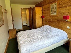Een slaapkamer met een houten hoofdbord, een wit geborduurde bedspreidek, een groen vloerkleed en een houten garderobe met een rood object bovenop.
