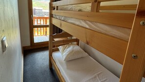 A bunk bed with a wooden headboard, a small window with a view of a balcony and mountains, and a door leading to an outdoor area.