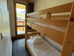 Un lit superposé avec une tête de lit en bois, une petite fenêtre donnant sur un balcon et des montagnes, et une porte menant à un espace extérieur.