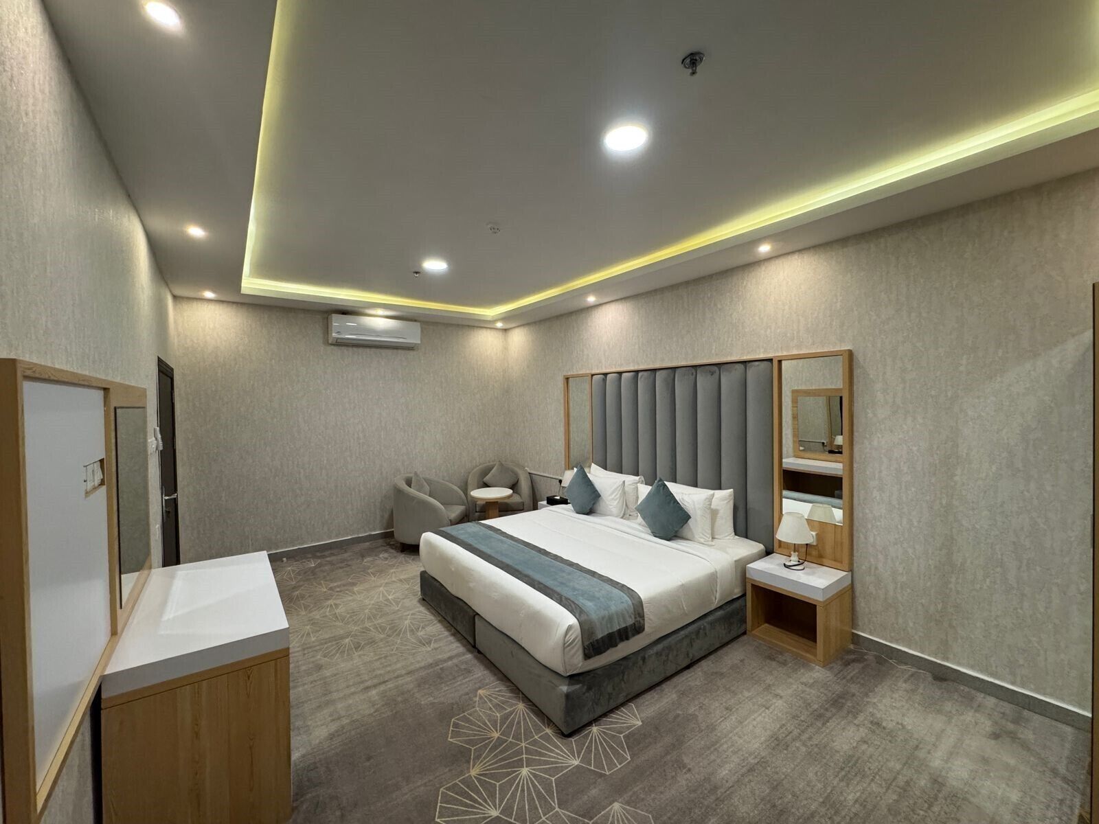 A modern hotel room with a large bed, bedside tables, a mirror, and a seating area with chairs.