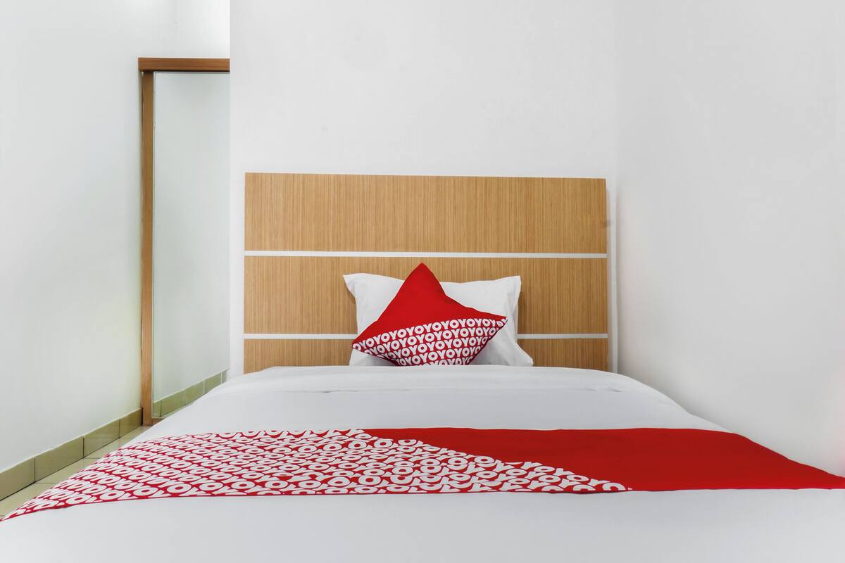 Una habitación de hotel con cabecera de madera, ropa de cama blanca y una manta decorativa con estampado rojo y blanco.
