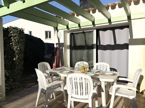 Eine überdachte Terrasse mit Esstisch für vier Personen, umgeben von weißen Plastikstühlen.